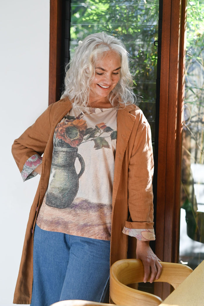 Woman wearing a floral t-shirt and brown coat standing indoors with a window in the background
