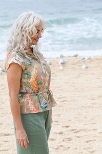 Woman standing on a beach wearing a floral sleeveless top and green pants.
